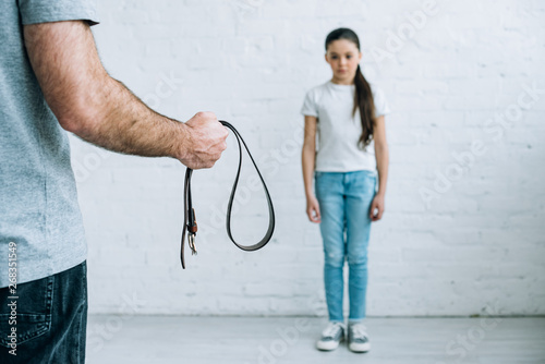 cropped view of father holding belt and upset daughter at home