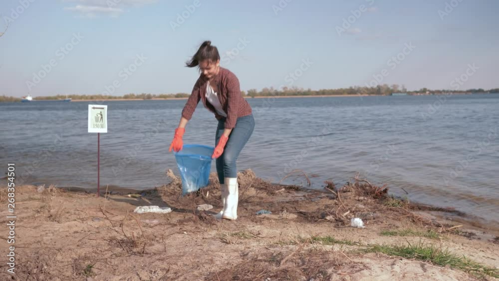 environmental solutions, happy girl volunteer collects plastic trash in garbage bag and gives positive gesture on polluted river beach near water