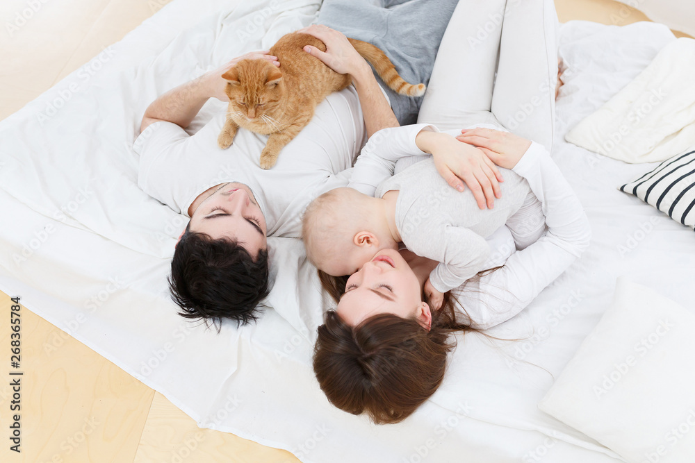 The family spends together time. Happy cute parents young girl and charming guy holding cute baby and red cat lying on sofa in home setting The family spends together time. Happy cute parents young girl and charming guy holding cute baby and red cat lying on sofa in home setting