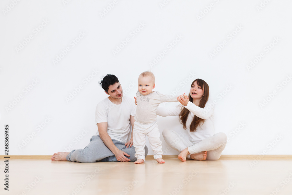 Naklejka premium Positive family portrait with mom,dad and little baby boy standing on white wall background. Parents hold by hand their child and help him do his first step. Generation concept