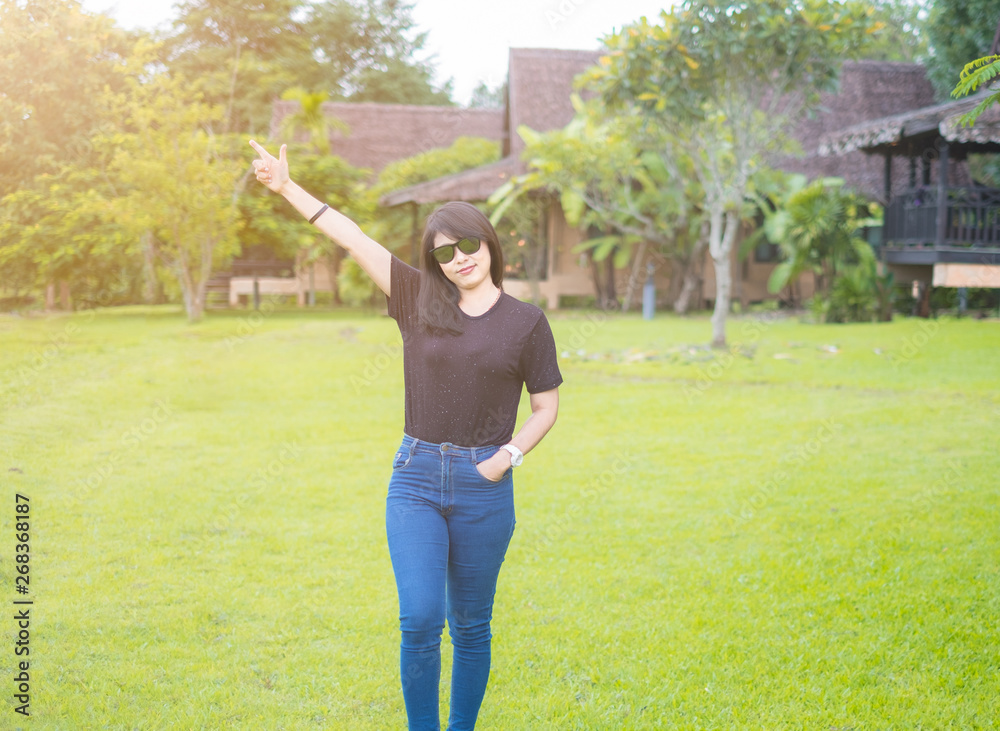 Beautiful Asian Women. wearing sunglasses. Standing poses lift Hands up In a good mood. Wear casual dress black t-shirt with Blue jeans. Green grass. enjoy relax Refreshing. sunlight of the morning.