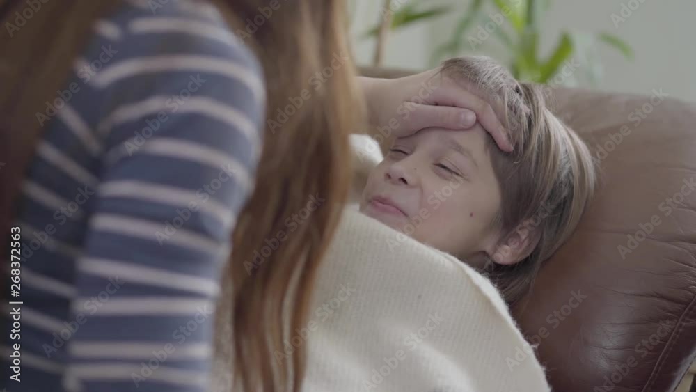 Small boy lie down in sofa under the blanket and girl touching his forehead with hand to see if brother have a temperature