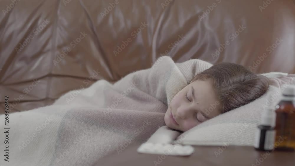 Teenage ill girl lying on the sofa covered with a blanket at home, she is cold. Nasal spray, pills and syrup on the table on the foreground. Concept of healthcare. Camera moving left Teenage ill girl lying on the sofa covered with a blanket at home, she is cold. Nasal spray, pills and syrup on the table on the foreground. Concept of healthcare. Camera moving left