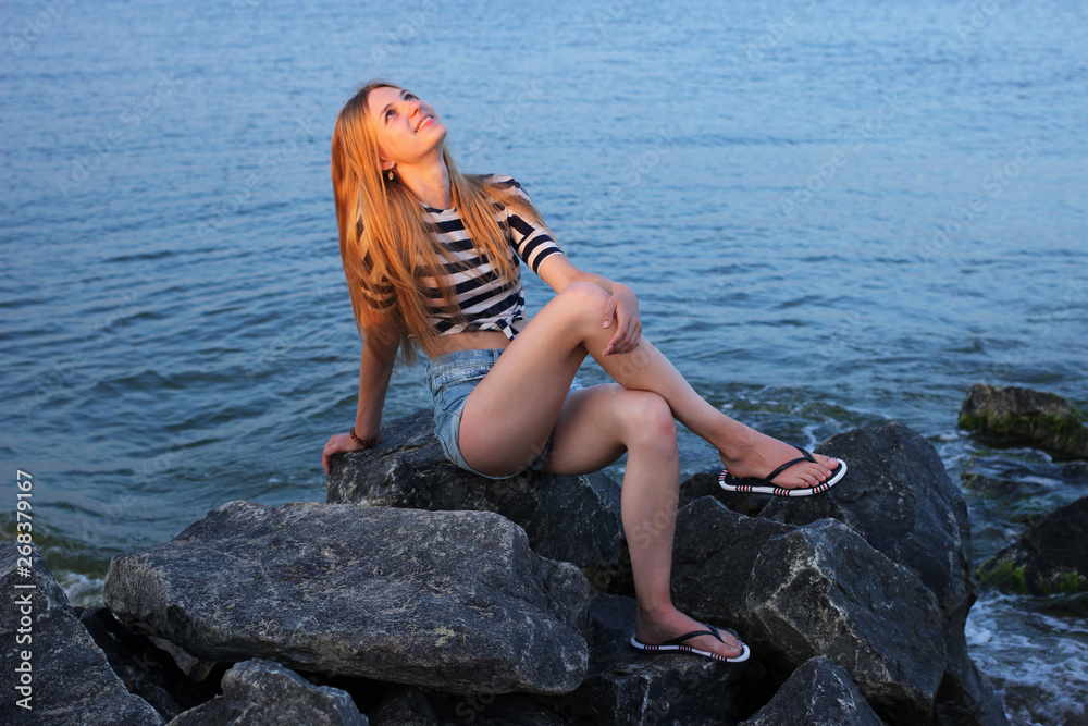 A charming girl sits on the rocks by the sea and admires the sunset.