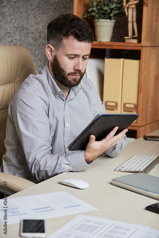 Bearded manager sitting at the table and working online using digital tablet at office Bearded manager sitting at the table and working online using digital tablet at office