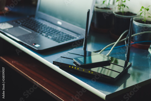 Black wireless with a smartphone on top and a laptop in background sitting on a desk in a small home office – Multiple modern portable gadget connected through Wi-Fi – Concept image for data sharing