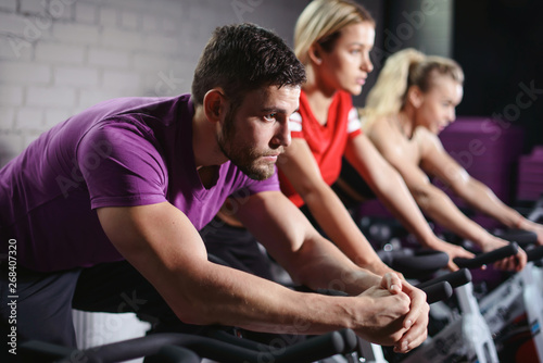 Close up hands of man biking in spinning class. Group of smiling friends at gym exercising on stationary bike. Happy cheerful athletes training on exercise bike.