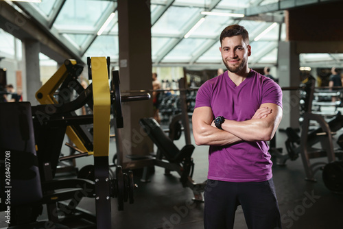 Handsome friendly personal trainer in gym. Young sporty man smiling standing on fitness club background