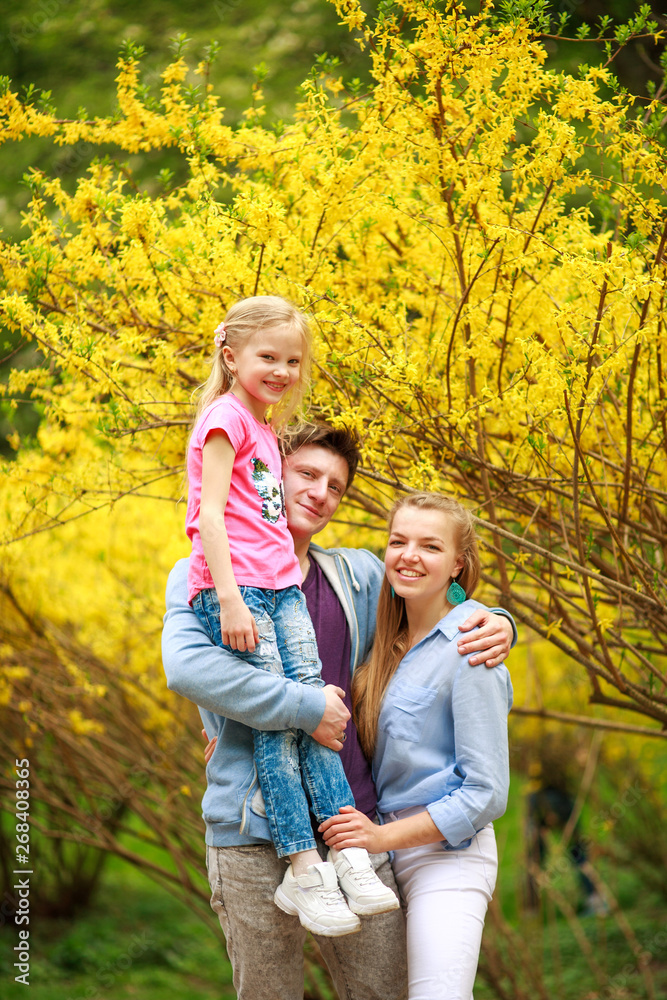 Young loving couple of parents with child daughter in park on background of yellow flowering tree Young loving couple of parents with child daughter in park on background of yellow flowering tree