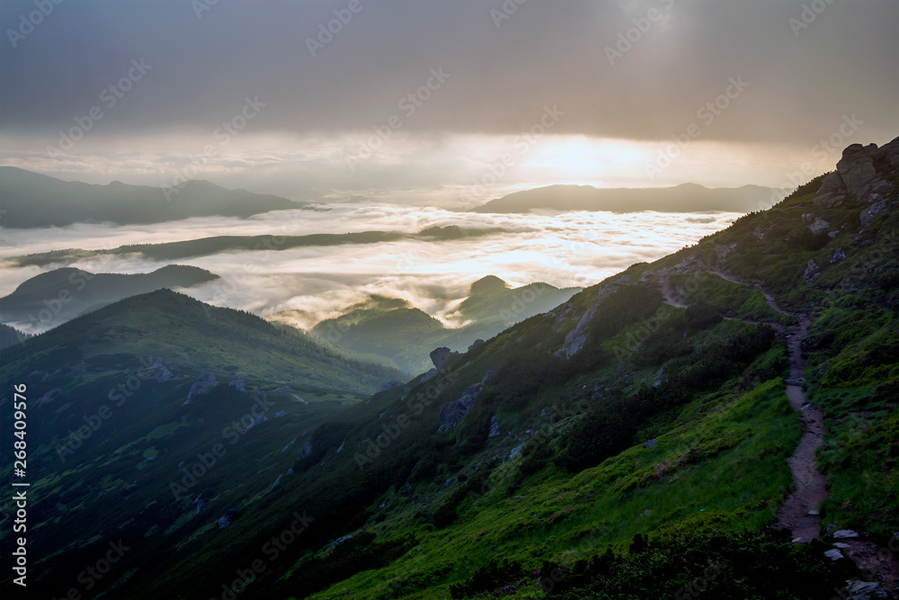 Obraz premium Fantastic view of mountain valley covered with low white puffy like snow clouds stretching to foggy horizon under bright morning sky with light orange glow at sunrise. Beauty of nature concept.