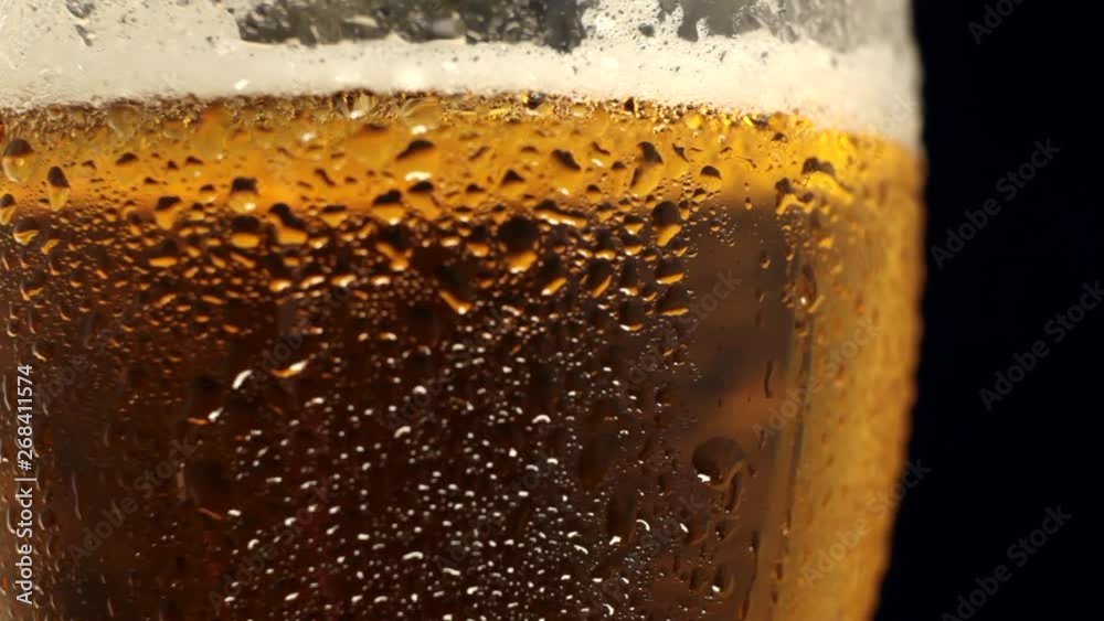 Glass of cold beer slowly rotates around its axis. Close-up of cold glass with water drops. Cold light beer with bubbles in a glass and dripping condensate drops. Glass of cold beer slowly rotates around its axis. Close-up of cold glass with water drops. Cold light beer with bubbles in a glass and dripping condensate drops.