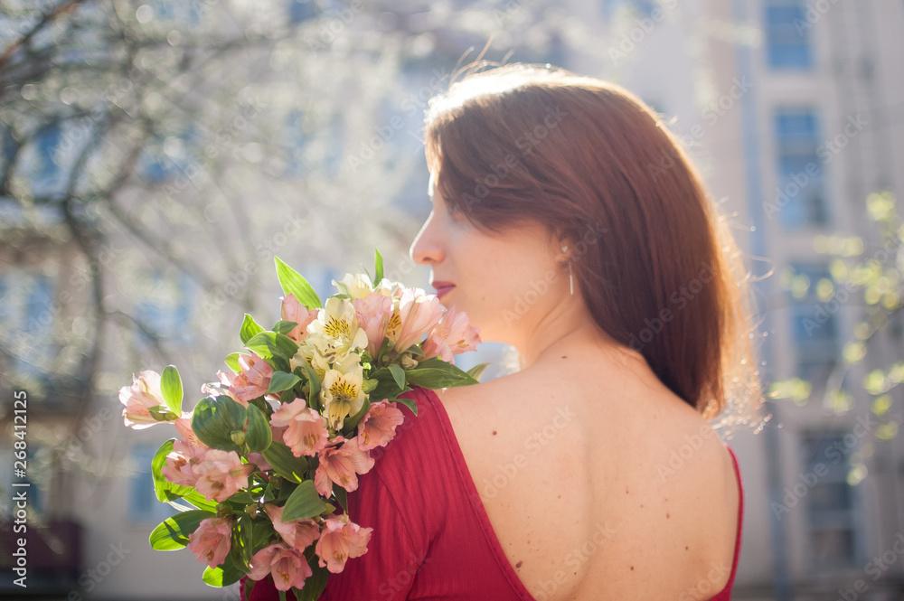Fototapeta premium Back view of amazing and beautiful young woman holding a big bouquet of colorful flowers outdoors near the buildings on the background