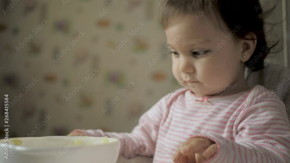Cute baby gil sitting in feeding chair and eating healthy food on kitchen Cute baby gil sitting in feeding chair and eating healthy food on kitchen