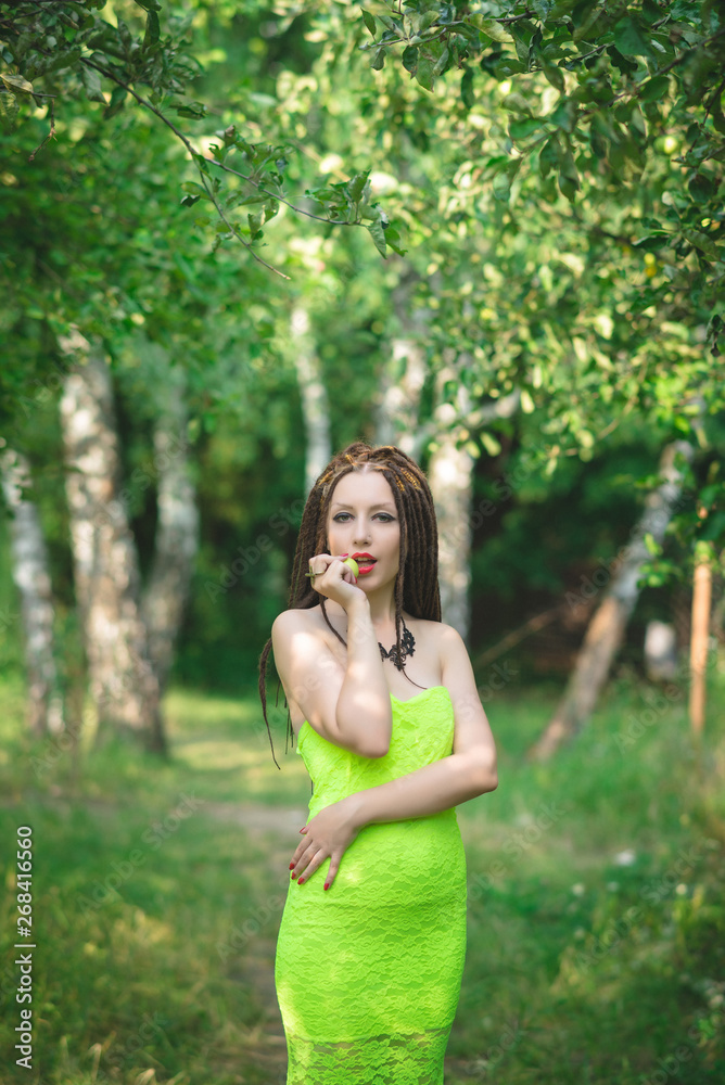 Barefoot girl with dreadlocks in a long green dress eating a green apple in the garden among the apple trees Barefoot girl with dreadlocks in a long green dress eating a green apple in the garden among the apple trees