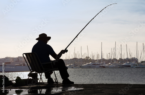 A patient angler is sitting in a chair at the pier of a Spanish port, hoping to catch a fish soon. In the background you can see sailboats in a marina.