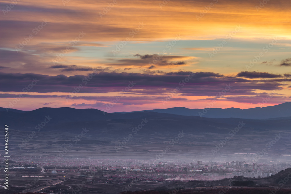 Dramatic, colorful sunset above distant city covered in mist and silhouette horizon mountains