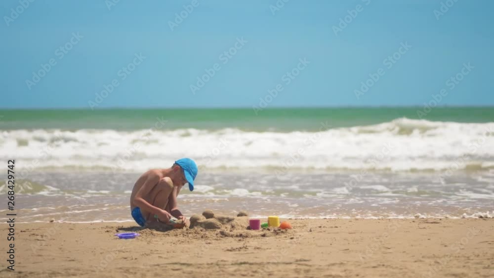 A little boy is playing in the sand on the sea, little legs and fingers, a background of sea yellow sand and blue water. Concept: children, childhood, summer, freedom, children, child. A little boy is playing in the sand on the sea, little legs and fingers, a background of sea yellow sand and blue water. Concept: children, childhood, summer, freedom, children, child.