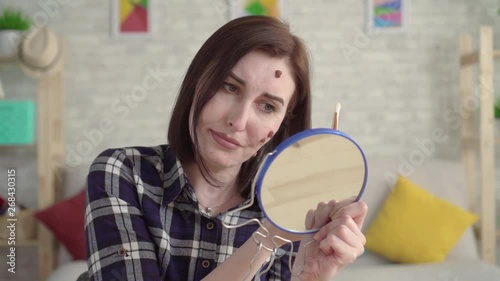 Young frustrated woman with moles on her face looking in the mirror and doing makeup close up photo