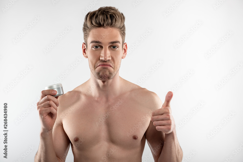 Handsome young man holding jar of cosmetic cream and showing thumbs up Handsome young man holding jar of cosmetic cream and showing thumbs up