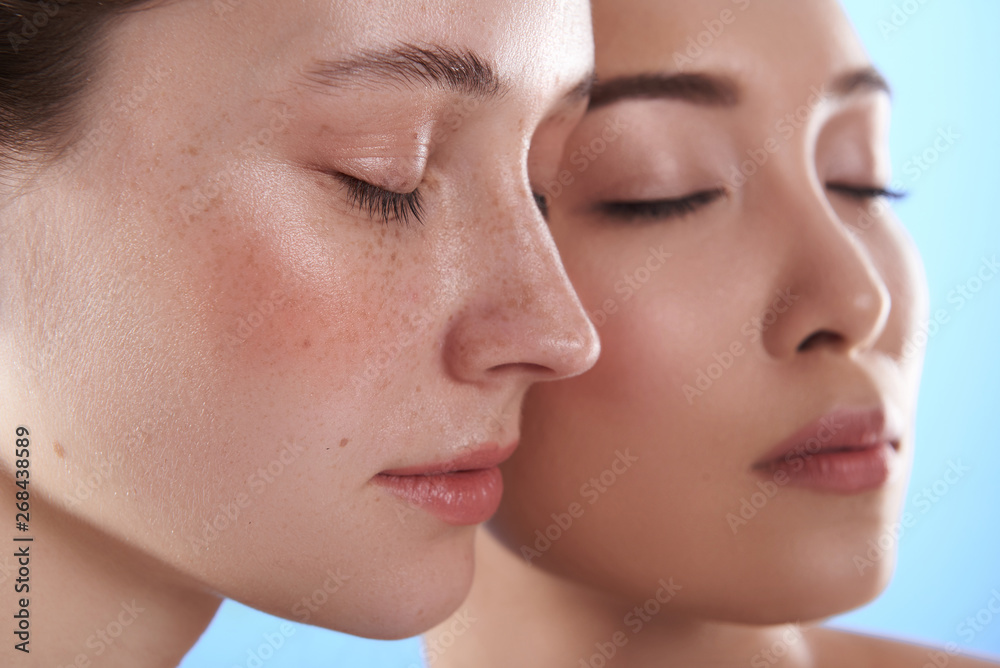 Close up of two interracial pretty women situating against blue background Close up of two interracial pretty women situating against blue background