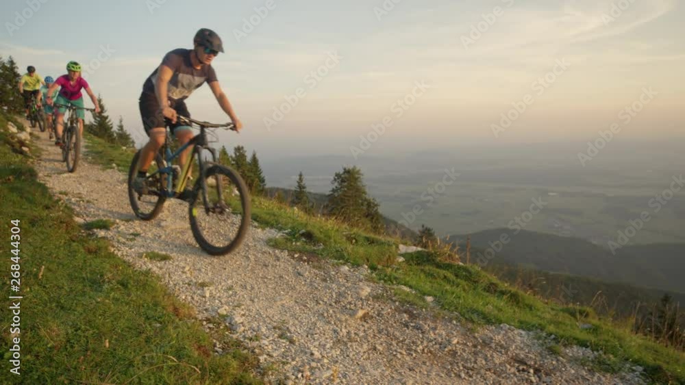 SLOW MOTION, CLOSE UP: Young mountain bikers ride along a narrow gravel path on a sunny summer morning. Four active friends having fun cross country biking in the idyllic green Slovenian mountains. SLOW MOTION, CLOSE UP: Young mountain bikers ride along a narrow gravel path on a sunny summer morning. Four active friends having fun cross country biking in the idyllic green Slovenian mountains.