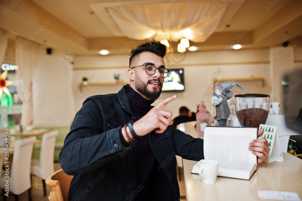 Arab man wear on black jeans jacket and eyeglasses in cafe drink coffee at bar with book. Stylish and fashionable arabian model guy. Arab man wear on black jeans jacket and eyeglasses in cafe drink coffee at bar with book. Stylish and fashionable arabian model guy.