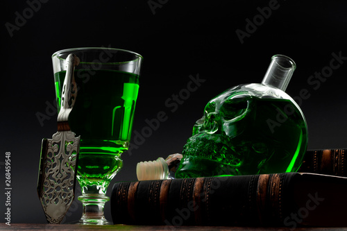 Alcoholic drink, creative stimulant and bohemian lifestyle concept theme with glass of absinthe and stainless steel spoon next to a skull shaped green bottle on a stack of books with black background photo