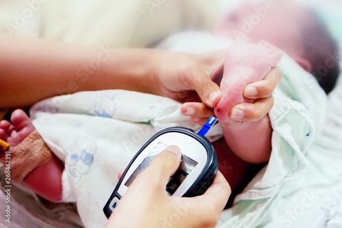 Hands of pediatric nurse hold and using blood and glucose meter with sheet check touch on sick newborn baby blood and foots to checkup glucose in his blood at NICU wards.