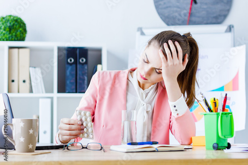 Young beautiful business woman boss feeling sick and tired  sitting at her workplace and taking pills. Exhausted girl employee with strong headache migraine is working in modern office.