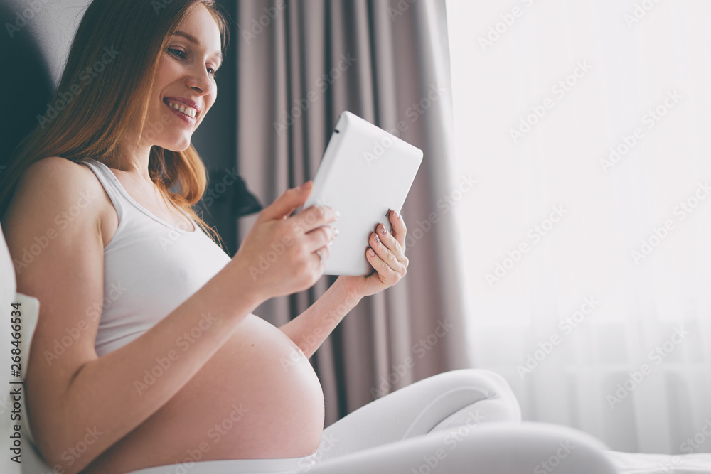 Technology and pregnancy. Young pregnant woman using tablet computer sitting on a bed at home. Technology and pregnancy. Young pregnant woman using tablet computer sitting on a bed at home.