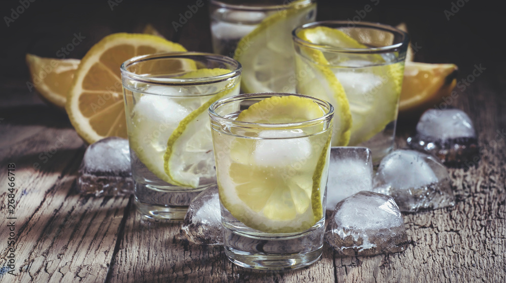 Lemon vodka with lemon slice and ice in shot glass, vintage wooden table, selective focus Lemon vodka with lemon slice and ice in shot glass, vintage wooden table, selective focus