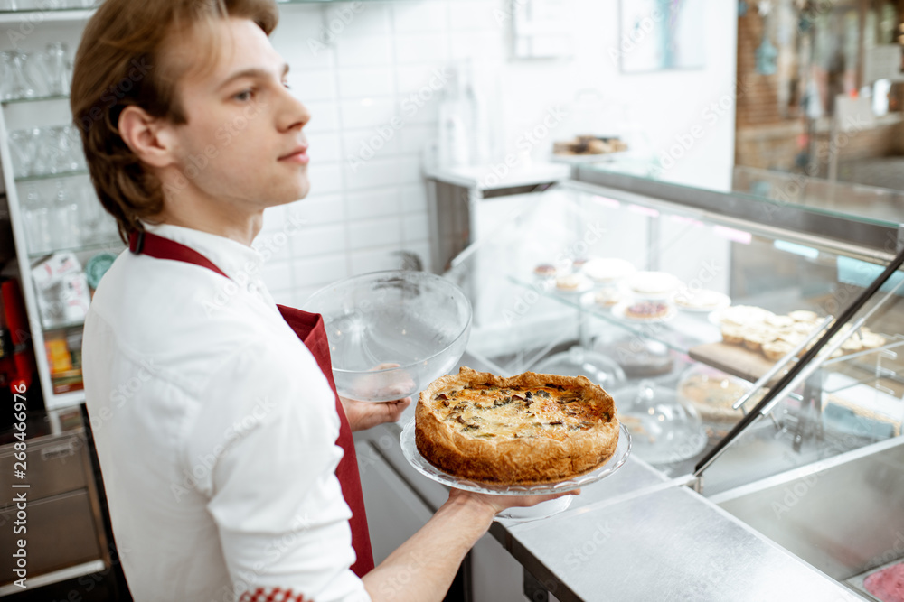 Salesman in red apron putting fresh pie into the refrigerator of the showcase at the modern pastry shop
