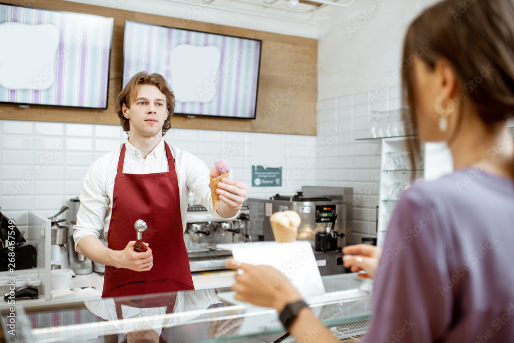 Handsome salesman in red apron making ice cream in waffle cone for a young woman client in the modern pastry shop Handsome salesman in red apron making ice cream in waffle cone for a young woman client in the modern pastry shop