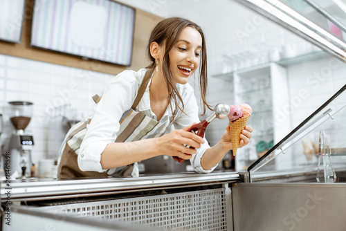 Young and happy saleswoman in apron making ice cream at the counter of the modern pastry shop indoors