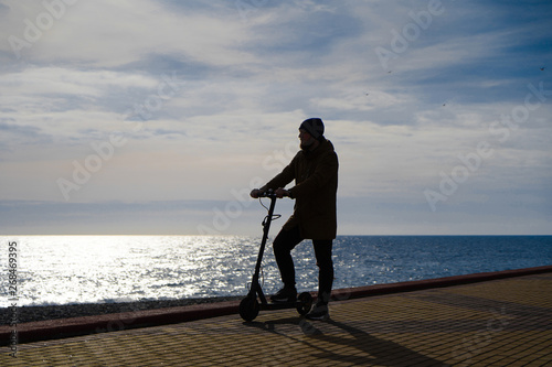 Adult man on an electric scooter on the embankment near the sea at sunset, looking into the distance, silhouette, free space © Ekaterina