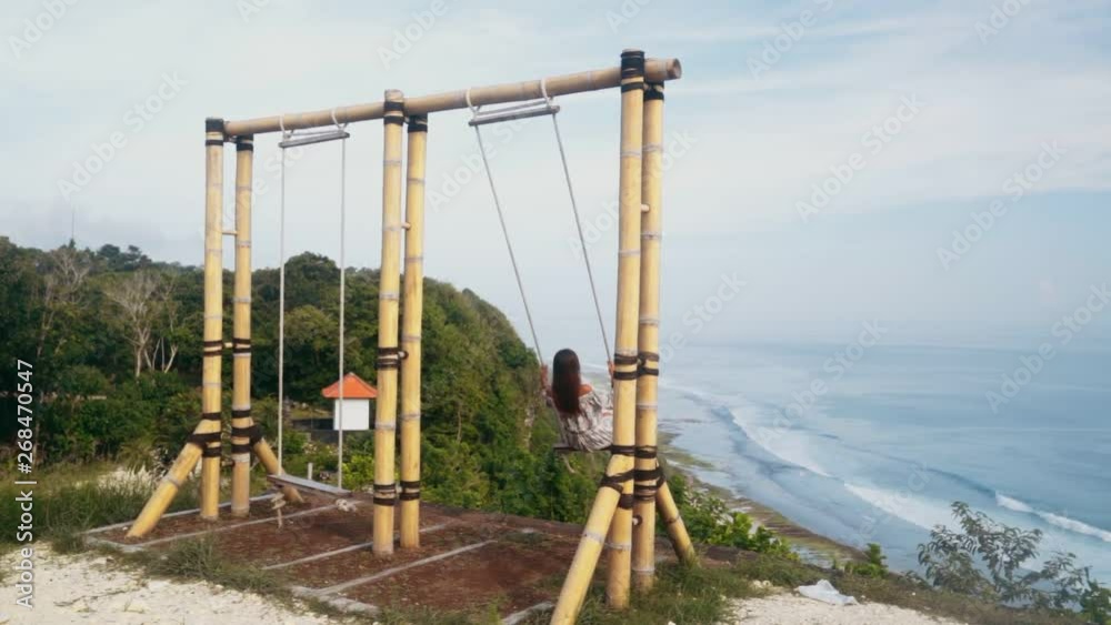 Backside view of young woman swings on swing with beautiful view on ocean, slow motion Backside view of young woman swings on swing with beautiful view on ocean, slow motion