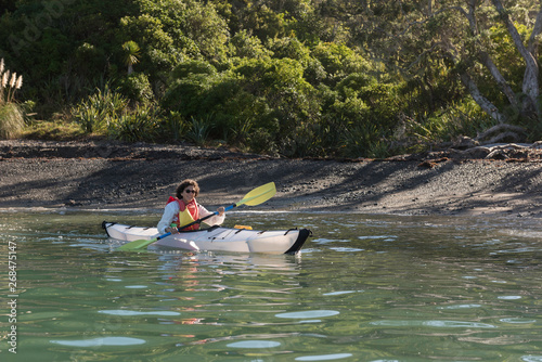 Female, baby boomer paddling her kayak on a sunny day by a beach on the calm waters of Whangaruru Harbour, Northland, New Zealand.