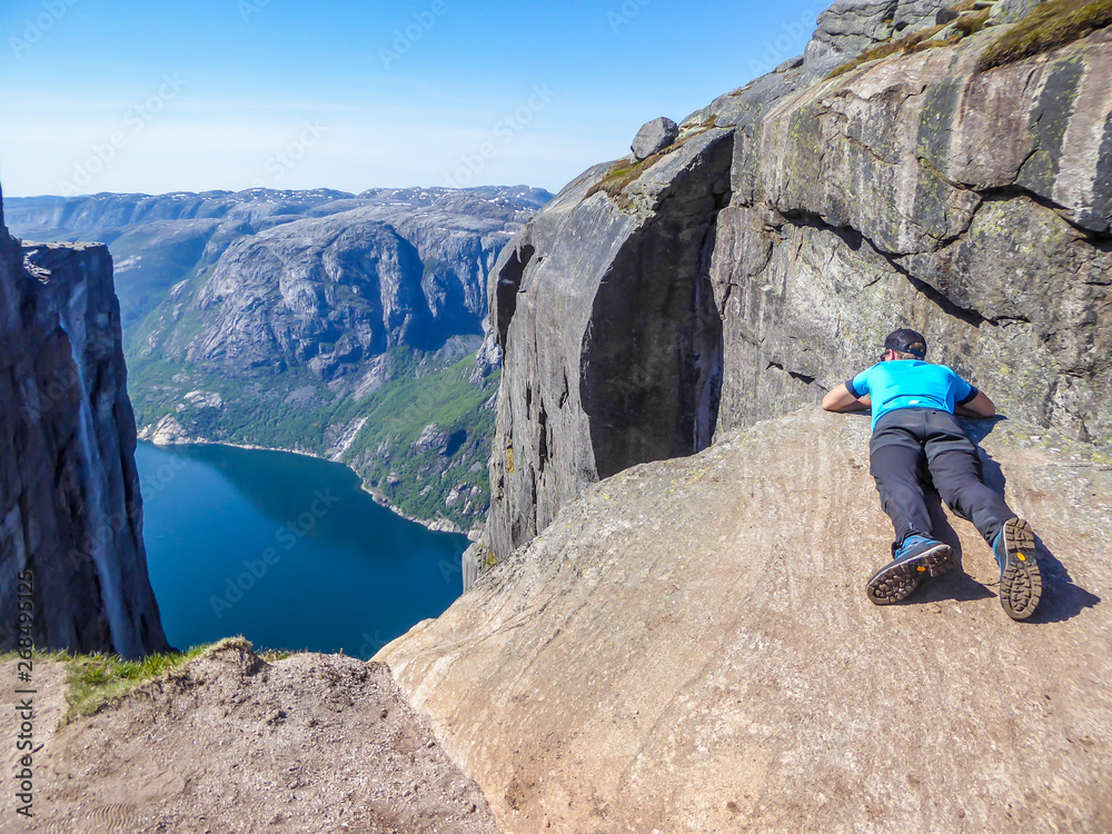 A young man lying at the edge of a steep mountain. He looks down into the valley. Overcoming the fear of heights. In front of her stunning Lysefjorden shimmering with many shades of blue and green