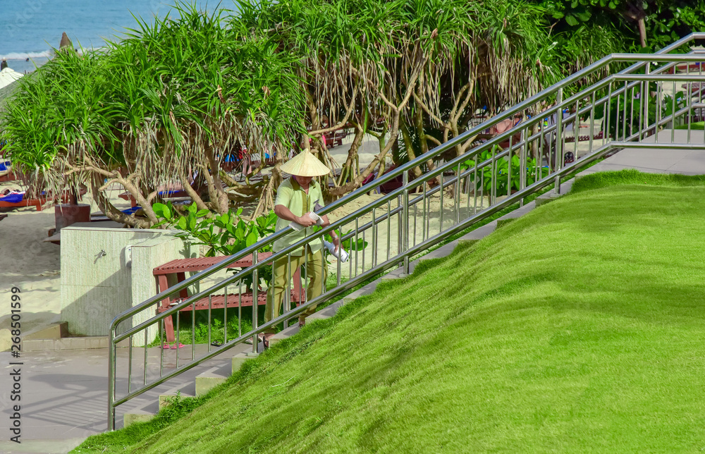 An uniformed employee does the cleaning on site. Rubs the chrome railing of the stairs. He is wearing a bamboo hat.