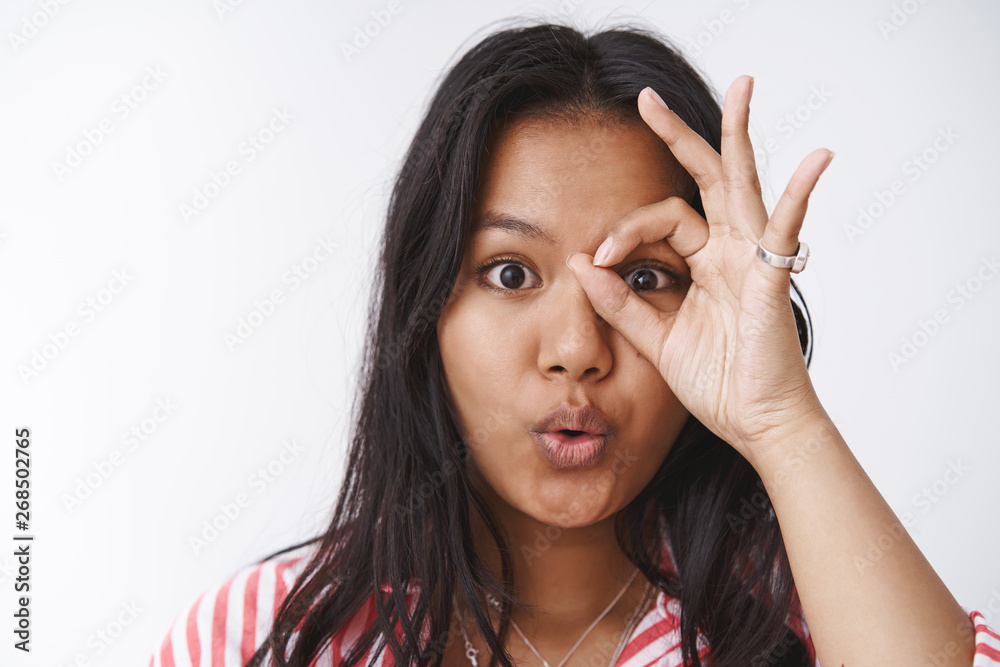 Close-up shot of intrigued and amazed enthusiastic young cute plump girl with tattoo showing ok or okay gesture over eye as peeking through hole at awesome thing, saying wow, folding lips curiously