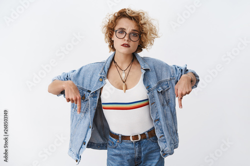 Questioned clueless cute young woman with short blond hair in transparent glasses and denim jacker pointing down with index fingers and looking unsure, hesitant at camera as asking question photo