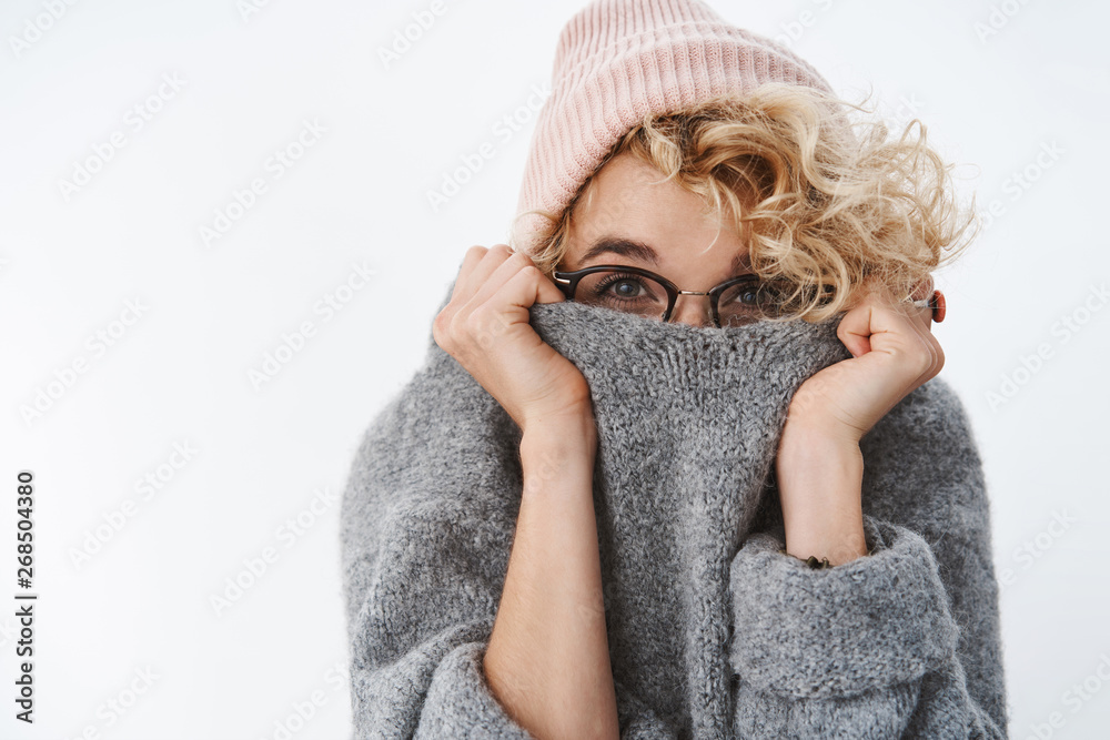 You cannot get me winter. Cute silly and playful blond girl in glasses and beanie pulling collar of sweater on face hiding and peeking at camera joyfully, having fun fooling around over white wall You cannot get me winter. Cute silly and playful blond girl in glasses and beanie pulling collar of sweater on face hiding and peeking at camera joyfully, having fun fooling around over white wall