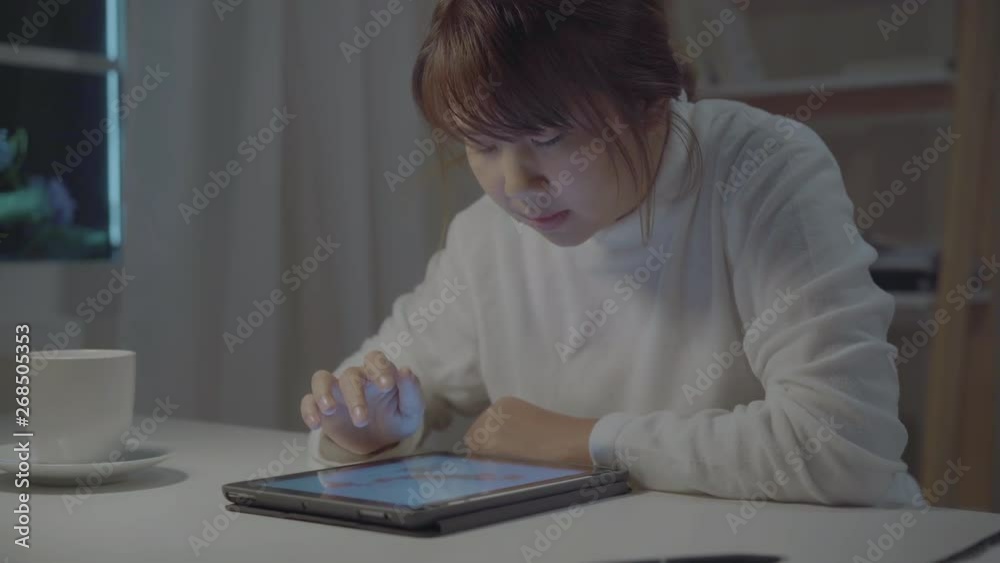Young asian woman working late using tablet checking social media while relax on desk in night in living room at home. Enjoying time at home concept. Young asian woman working late using tablet checking social media while relax on desk in night in living room at home. Enjoying time at home concept.