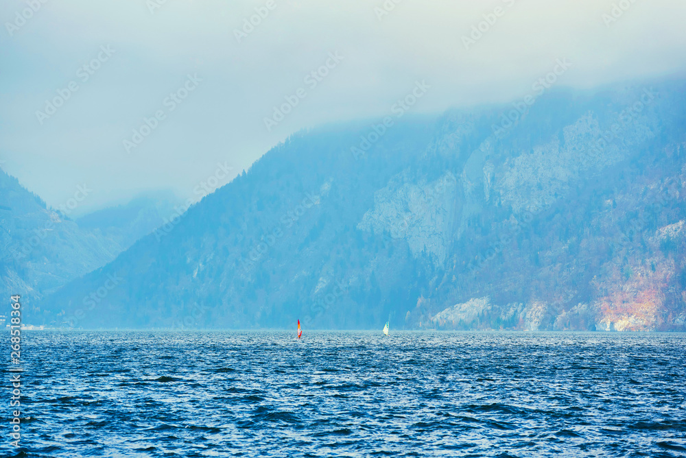 Windsurfing on lake landscape and foggy mountains at background. Man Windsurfer on the Board with a sail floating on the lake.