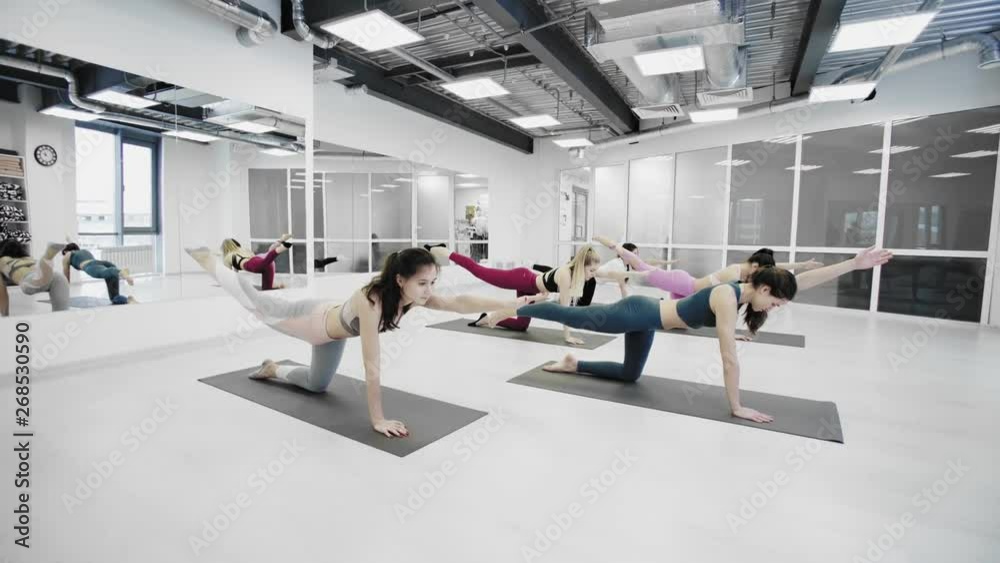 Sporty attractive girls are doing stretching exercises forward bending head to knee position while sitting on colorful yoga mats in light wellness studio.