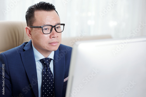 Serious Asian businessman in eyeglasses sitting in front of computer and looking at computer monitor at office
