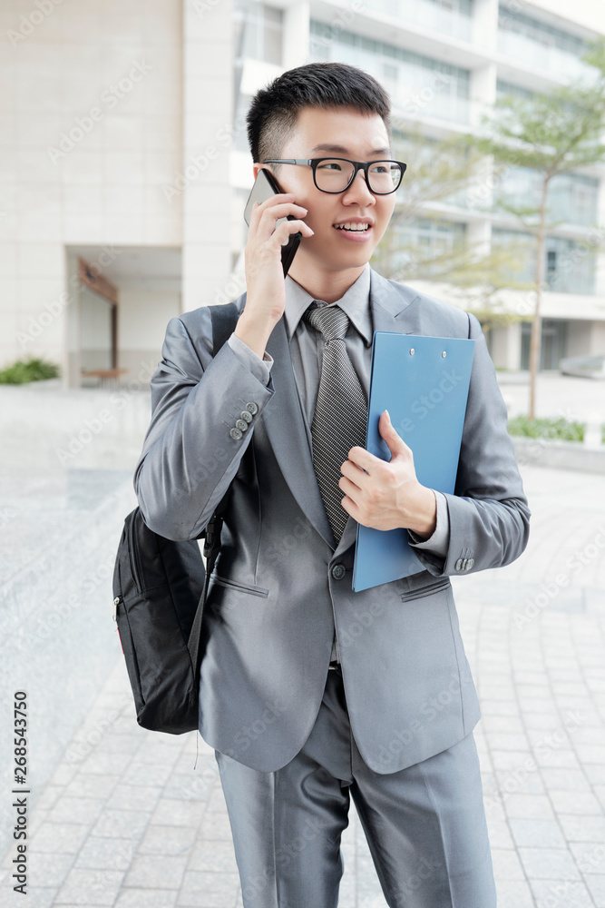 Asian young office worker holding clipboard and has a conversation on mobile phone while standing in the city Asian young office worker holding clipboard and has a conversation on mobile phone while standing in the city