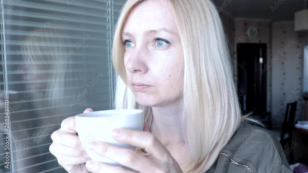 Portrait of young pretty attractive blond woman with blue eyes standing near the open window in the morning, drinking hot coffee and looking into the distance Portrait of young pretty attractive blond woman with blue eyes standing near the open window in the morning, drinking hot coffee and looking into the distance