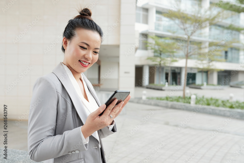 Asian young businesswoman looking at her mobile phone and smiling while standing in the city near the office building