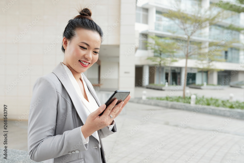 Asian young businesswoman looking at her mobile phone and smiling while standing in the city near the office building Asian young businesswoman looking at her mobile phone and smiling while standing in the city near the office building